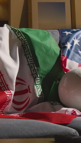 Vertical-Video-Of-Sofa-In-Lounge-With-Iranian-And-American-Flags-And-Ball-As-Fans-Prepare-To-Watch-Football-Soccer-Match-On-TV